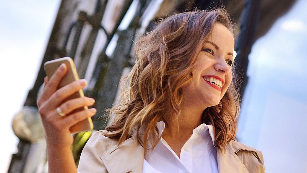 Una donna è in piedi davanti a una vetrina, sorride e tiene un cellulare nella mano destra.
